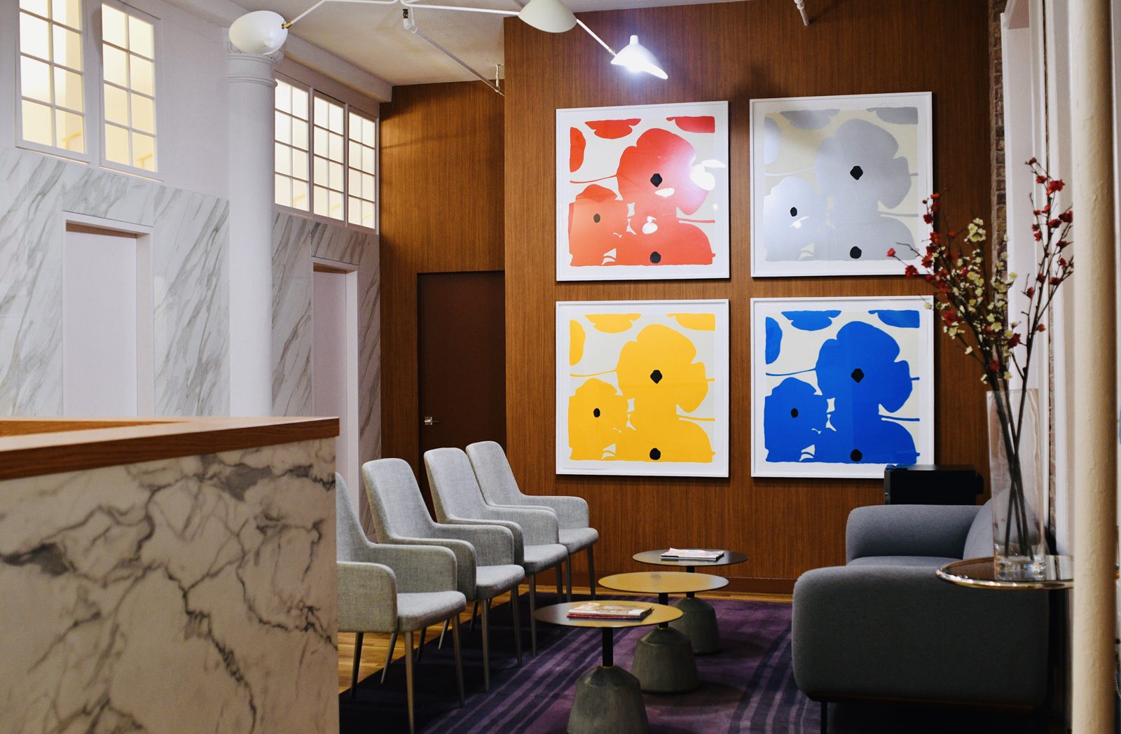 Soho Dermcare reception at 594 Broadway — Calacatta-marble desk, warm wood feature wall with four framed pop-art floral prints, grey upholstered chairs and tall loft windows